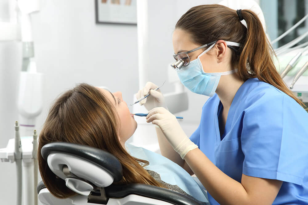 The image shows a dental professional wearing blue scrubs and gloves performing a dental procedure on a patient reclining in a dental chair. The practitioner is using dental instruments, working inside the patient's mouth. The setting is a dental clinic, with various dental equipment visible in the background, emphasizing a routine healthcare scenario focused on oral health.