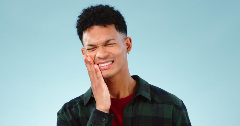 A man grimaces in pain, holding his jaw with one hand as he appears to suffer from severe discomfort. His expression and posture emphasize distress, likely due to a dental issue or injury.